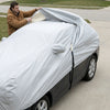 A man pulling the silver-grey car cover over the roof of a sedan on a driveway