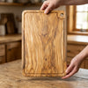 Two hands lifting the Alba Olive Wood Carving Board with Juice Groove off a wooden table