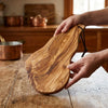 Two hands lifting the Alba Olive Wood Board with Leather Loop off a wooden table