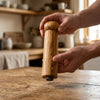 Two hands lifting the Alba Olive Wood Pepper Mill off a wooden table